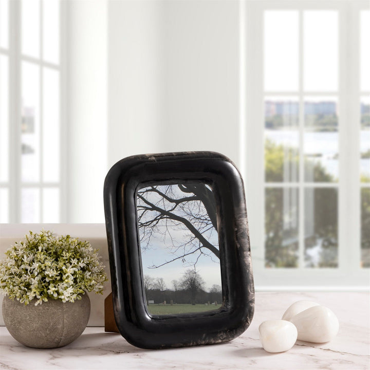 Black decorative picture frame on a table with a plant and stones in a bright room.