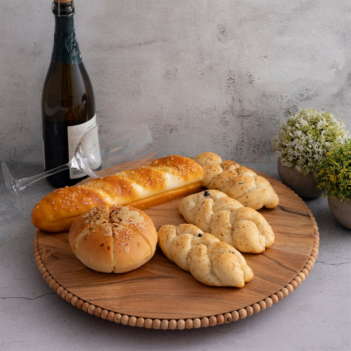 Assorted bread loaves on a wooden beaded plate with a bottle of wine and glasses against a gray background.