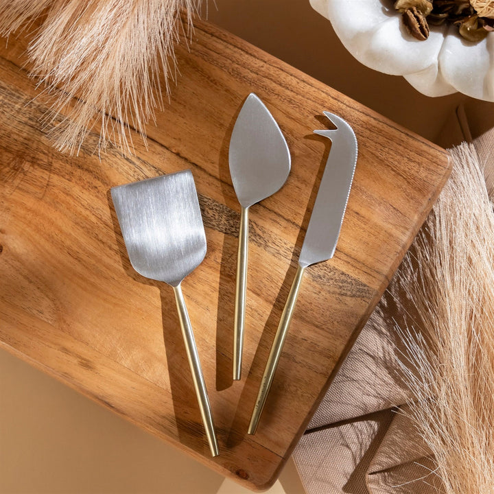 Three metal cheese knives on a wooden board with decorative elements.