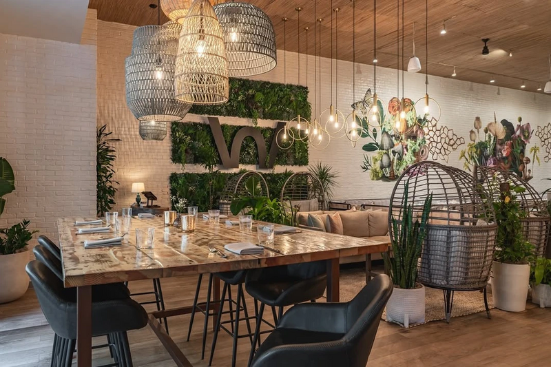 Communal restaurant table with woven pendants and a preserved greenery wall